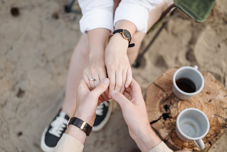 A couple's hands held gently, symbolizing love and connection. Outdoors with coffee cups and a rustic setting.