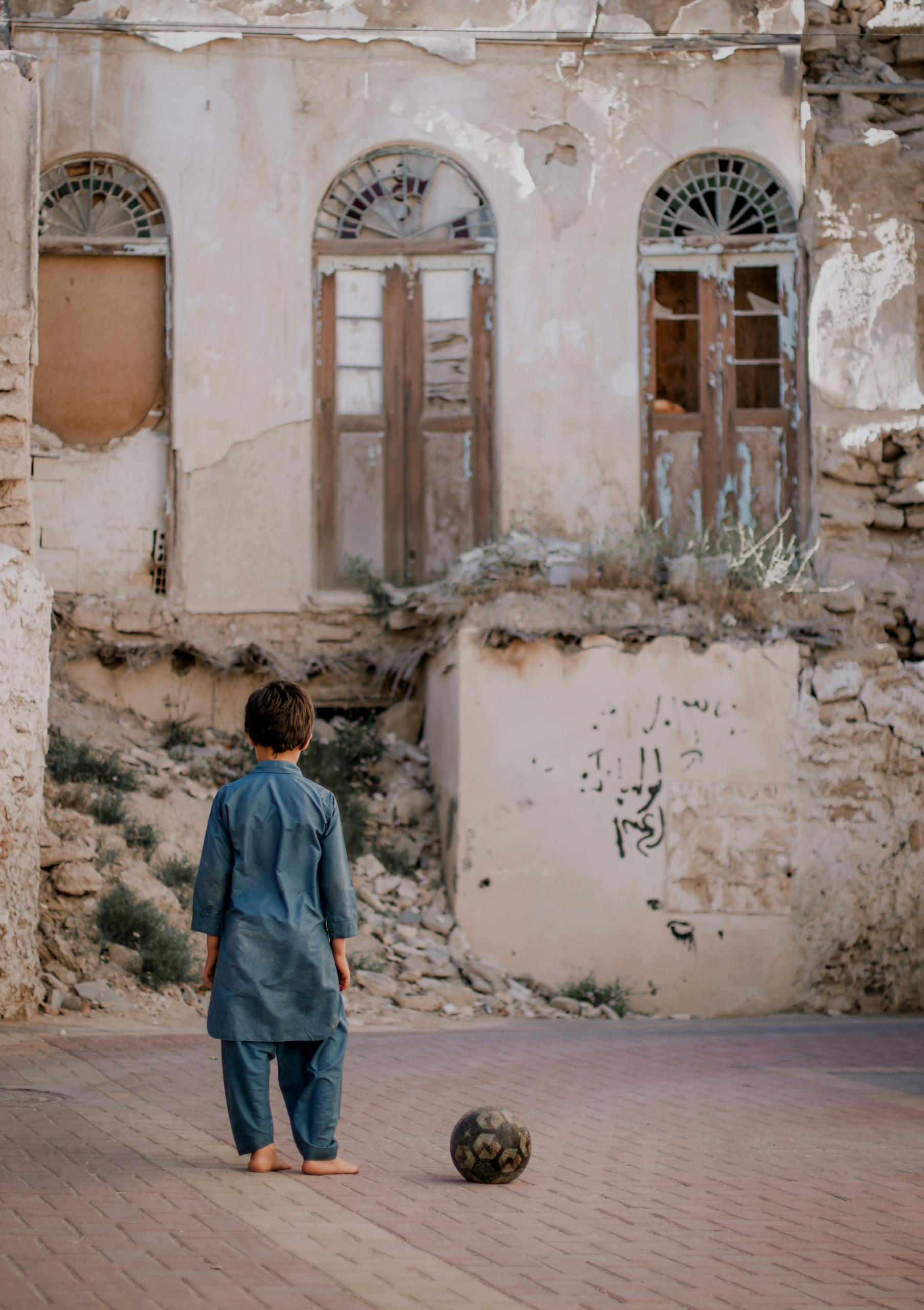A boy stands with a ball on a pavement in front of an abandoned building in Iran.