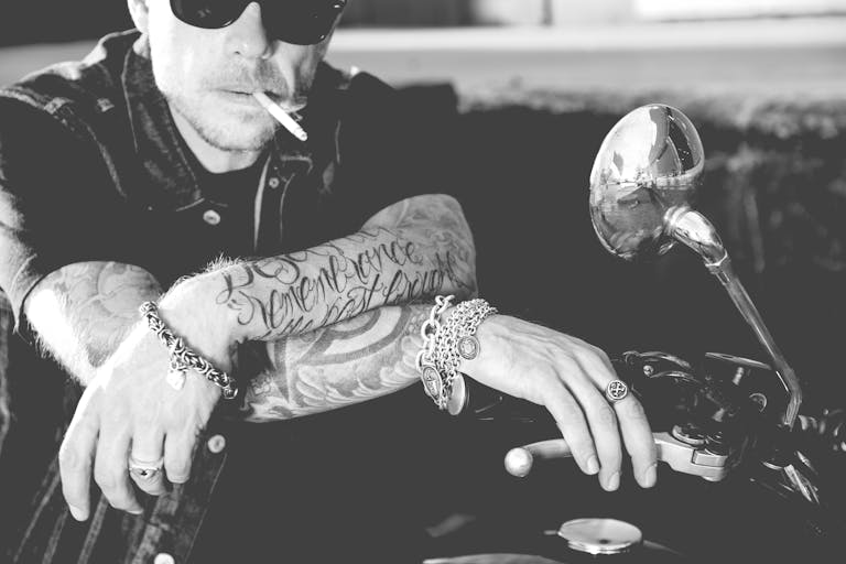 Black and white photo of a tattooed man with jewelry sitting on a motorbike, exuding individuality.