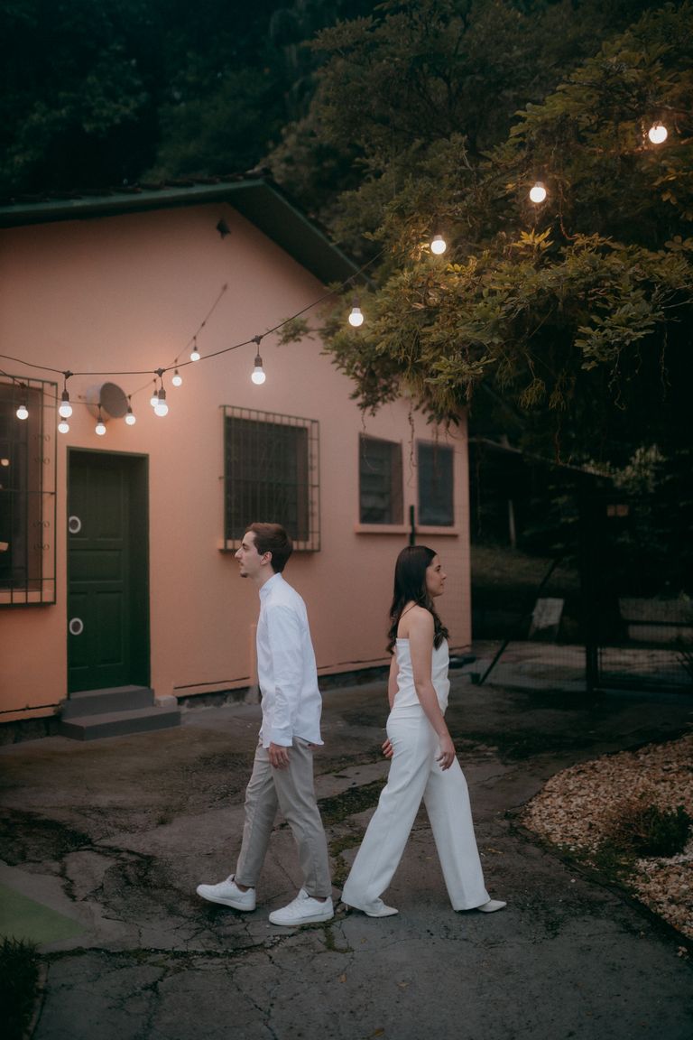 A couple walks together near a house adorned with string lights in a peaceful outdoor setting.