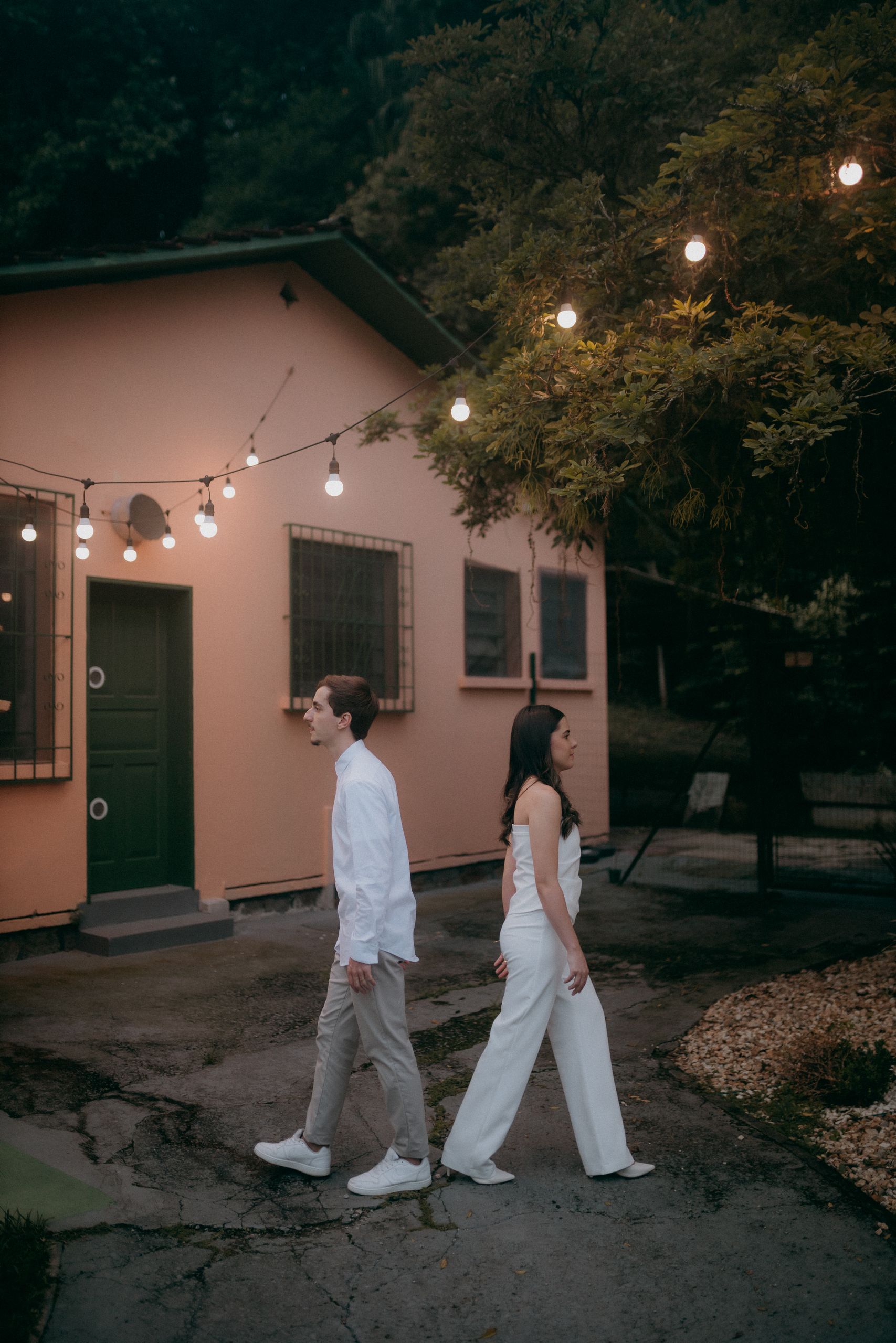 A couple walks together near a house adorned with string lights in a peaceful outdoor setting.