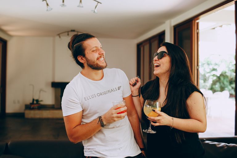 Cheerful young man and woman in trendy casual clothes standing near each other and laughing while drinking cocktails during meeting at home